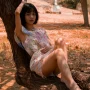 A woman in a floral dress leans on a tree branch in a sunny park, evoking leisure and tranquility.